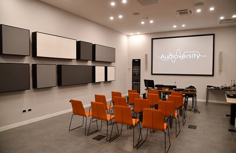 Lesmo training facility with orange chairs, presentation screen, and wall-mounted audio equipment.