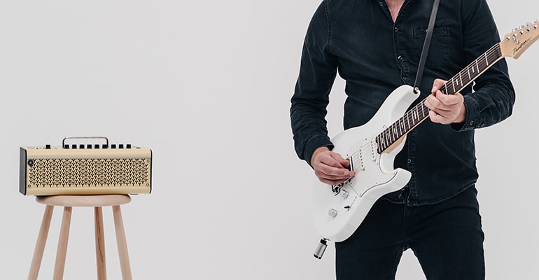 A man on a gray background plays the white Pacifica Standard Plus electric guitar that the YW10T is plugged into its jack. The THR30II Wireless guitar amp on a wooden stool is on the right behind the man. The guitar sound comes out from the THR30II Wireless through the YW10T.